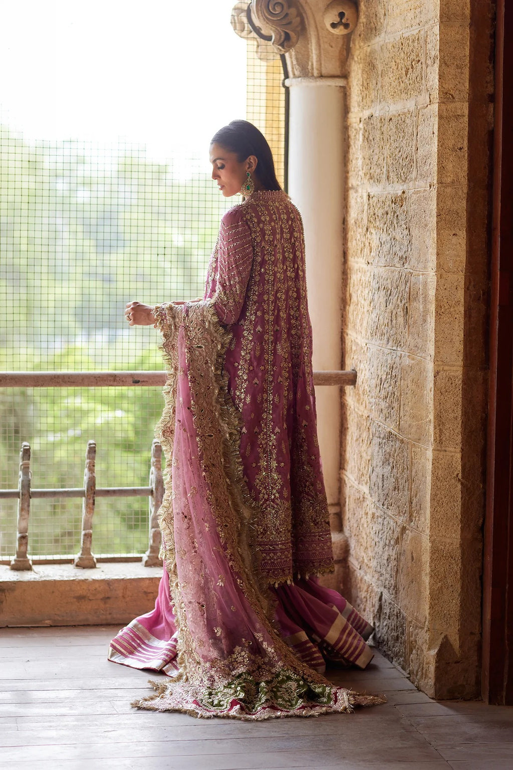 A woman wearing an ornate pink and gold embroidered traditional outfit stands on a stone balcony, holding out one of the wide sleeves. The background features a stone wall, a column, and a window looking out onto greenery.