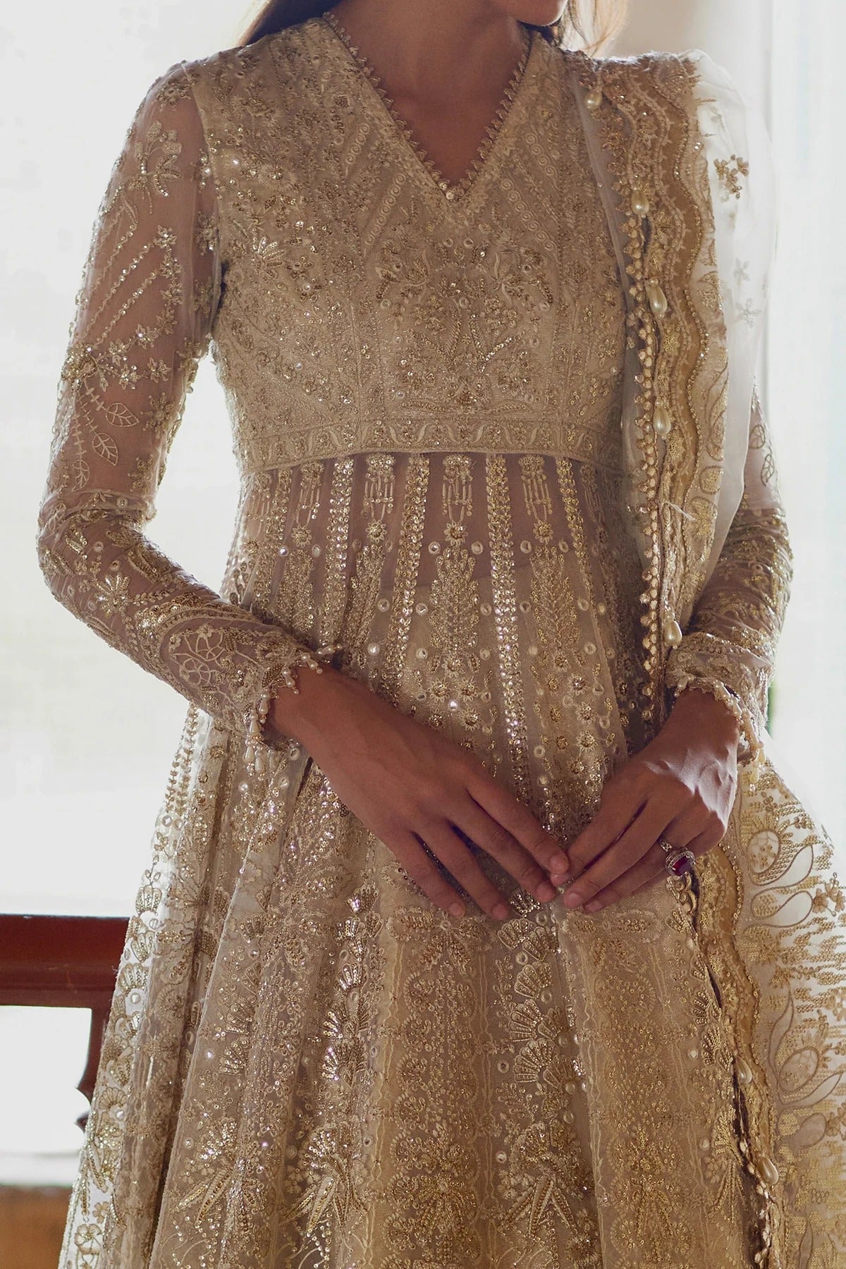 A close-up of a woman wearing a heavily embellished gold traditional dress with intricate embroidery, sheer long sleeves, and a matching embroidered dupatta draped over one shoulder.