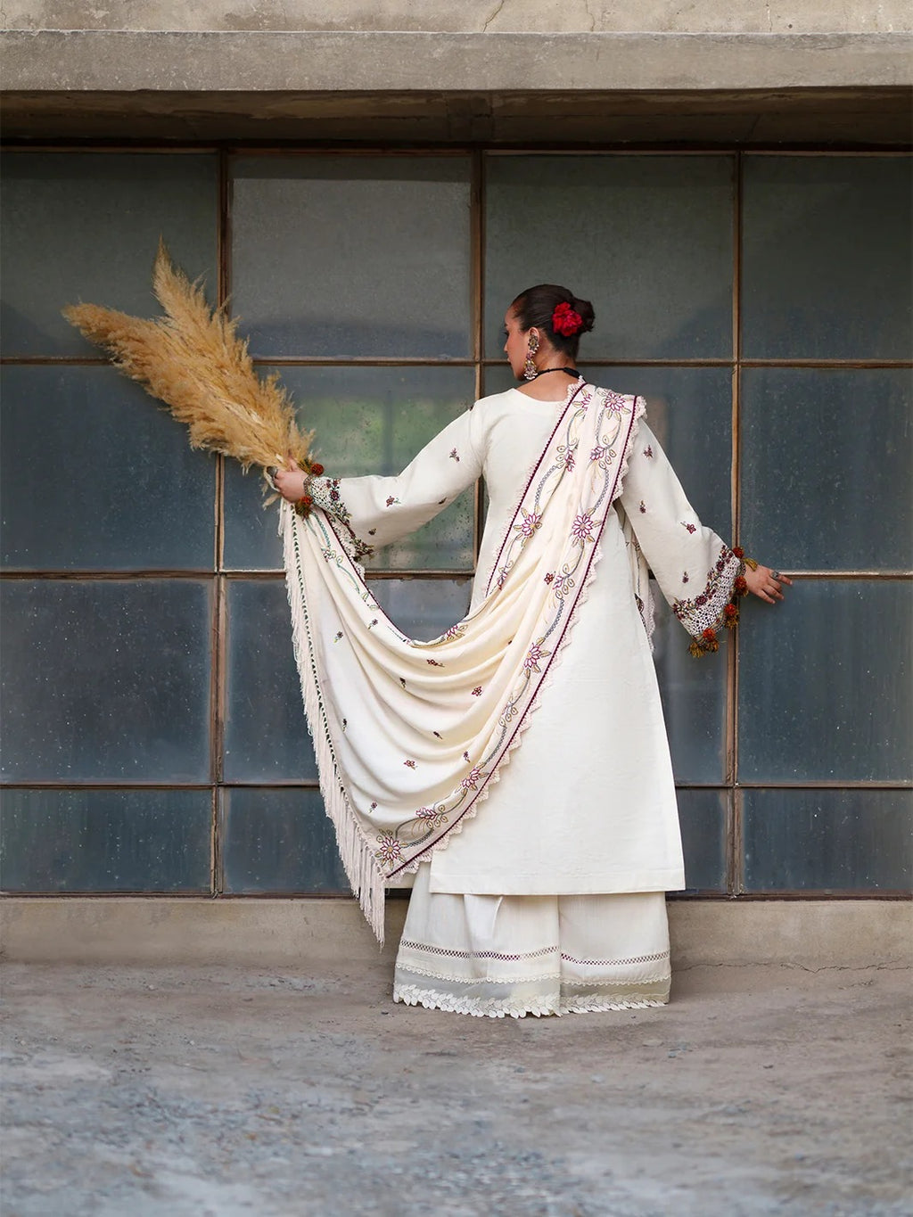 A woman stands with her back to the camera in front of a large paneled wall, wearing a cream-colored traditional outfit with embroidered details and holding a bundle of dried pampas grass.