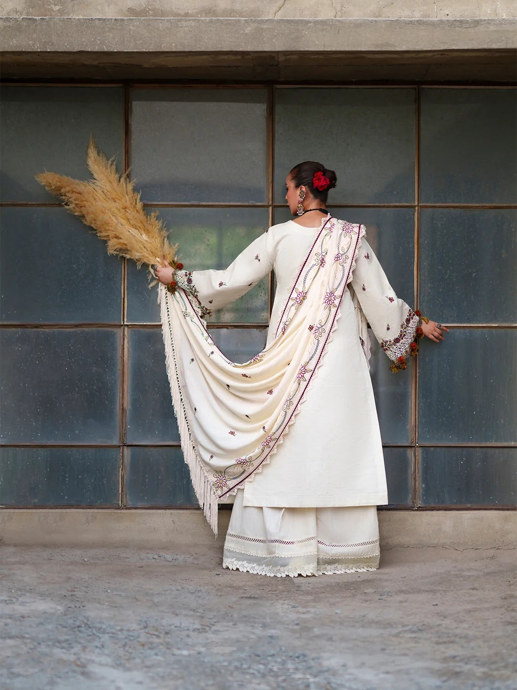 A woman stands with her back to the camera in front of a large paneled wall, wearing a cream-colored traditional outfit with embroidered details and holding a bundle of dried pampas grass.