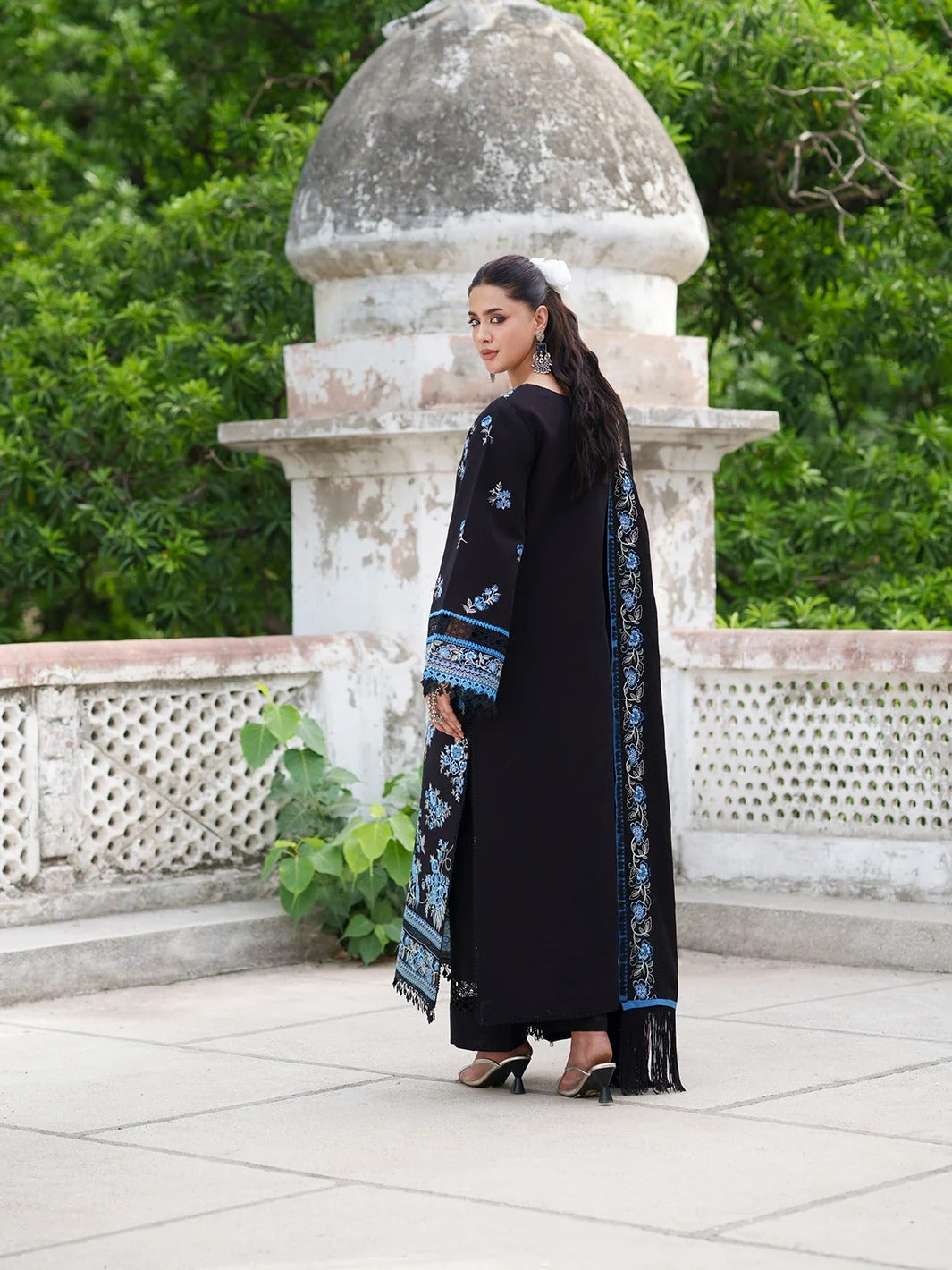 A woman wearing a black traditional outfit with blue and white embroidered details stands outdoors in front of a stone dome structure and green foliage.