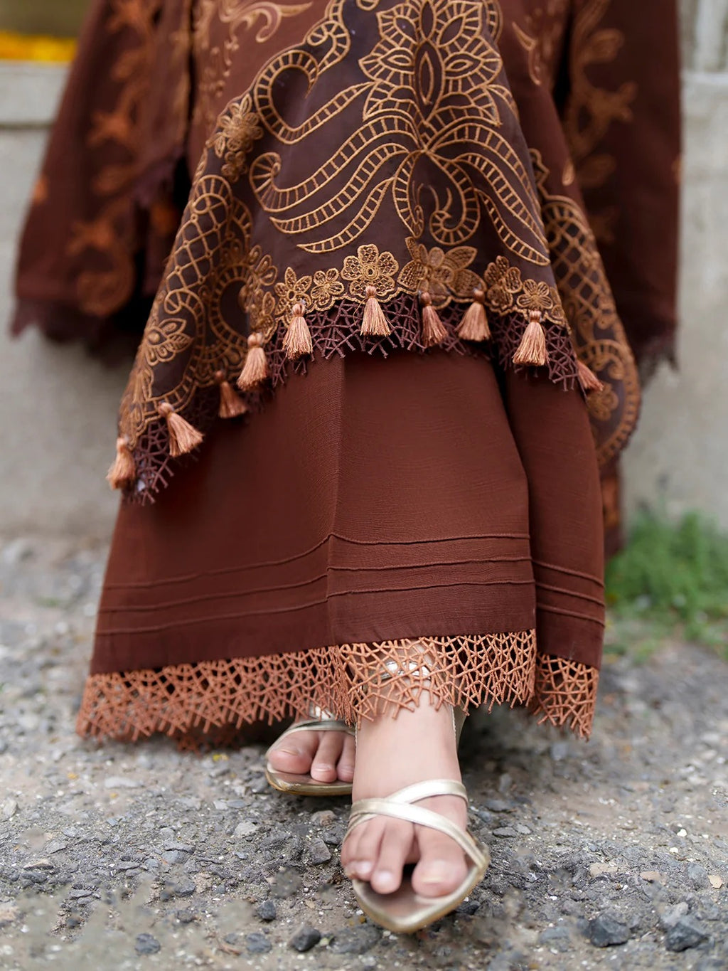 A close-up of a person wearing a brown traditional outfit with intricate gold embroidery, lace trim, and tassel details, paired with metallic gold flat sandals, standing outdoors on a gravel surface.
