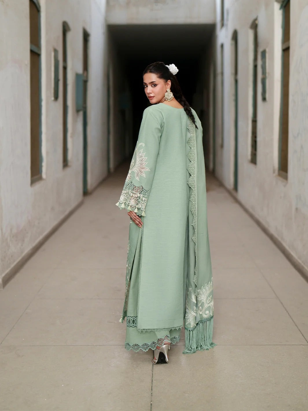 A woman stands in a long corridor wearing a sage green traditional outfit with white floral embroidery on the sleeves and hem, paired with a matching dupatta.