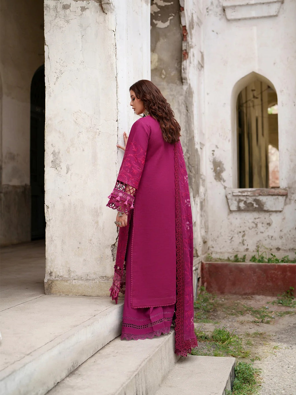 A woman wearing a magenta pink traditional South Asian outfit with matching dupatta and flared pants, standing on stone steps in front of a weathered white building.