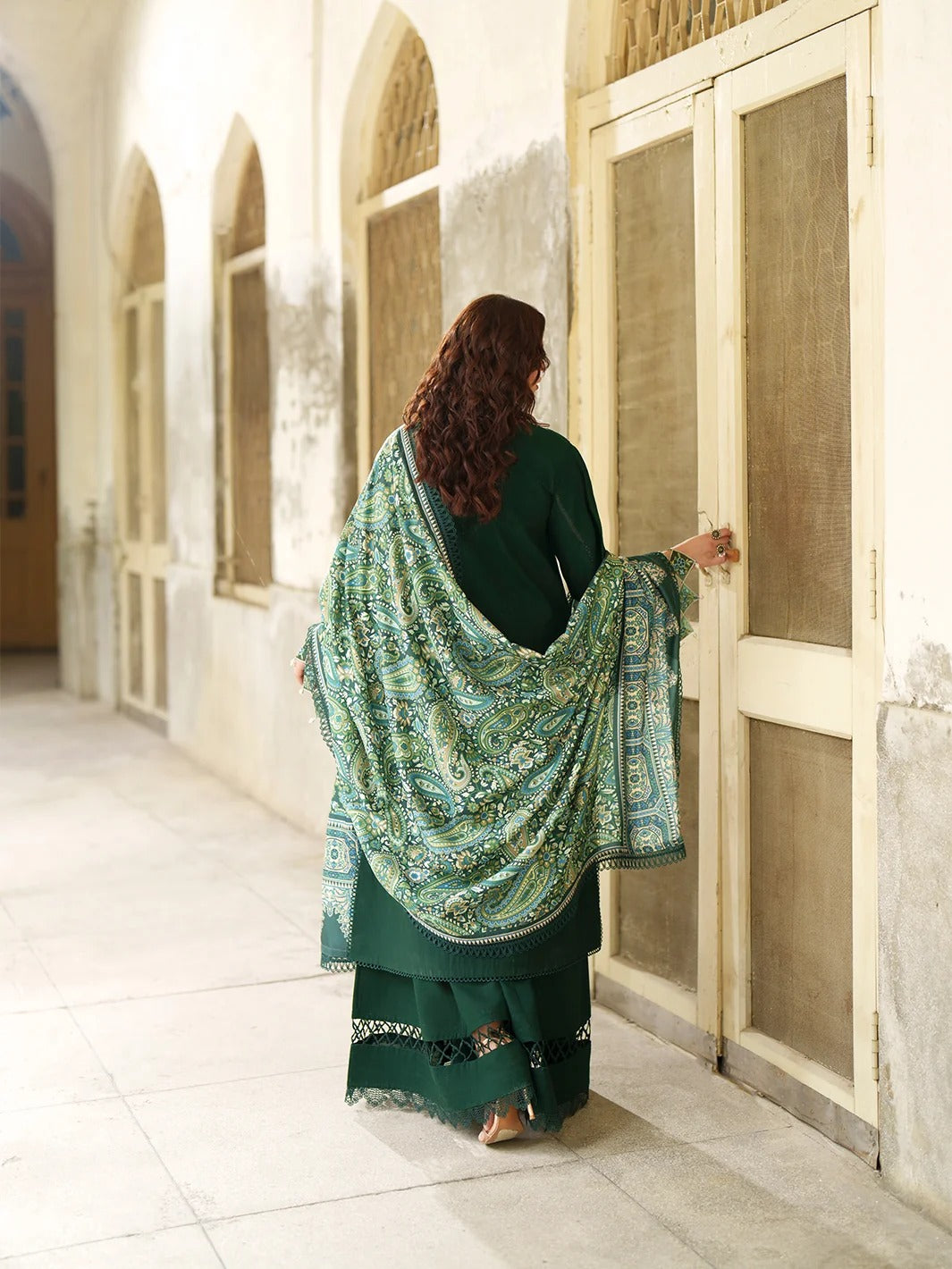 A woman in a dark green traditional outfit with a patterned green and blue shawl stands with her back to the camera, opening a large cream-colored wooden door in an arched corridor.