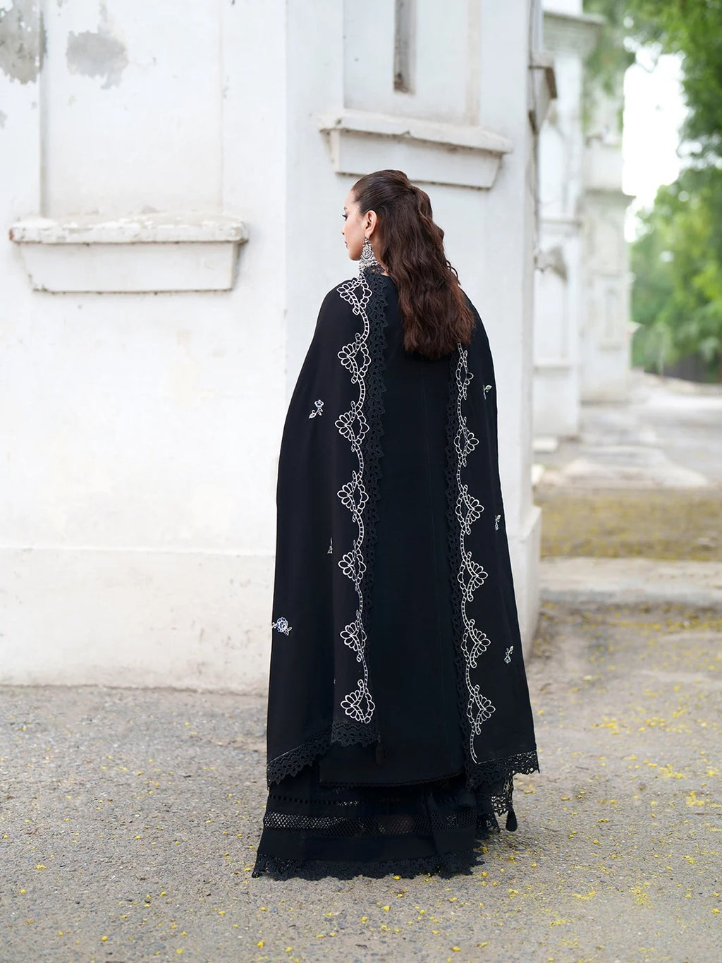 A woman stands outdoors in profile wearing a long black embroidered garment or drape with ornate silver-toned lace-like detailing running vertically along the edges.