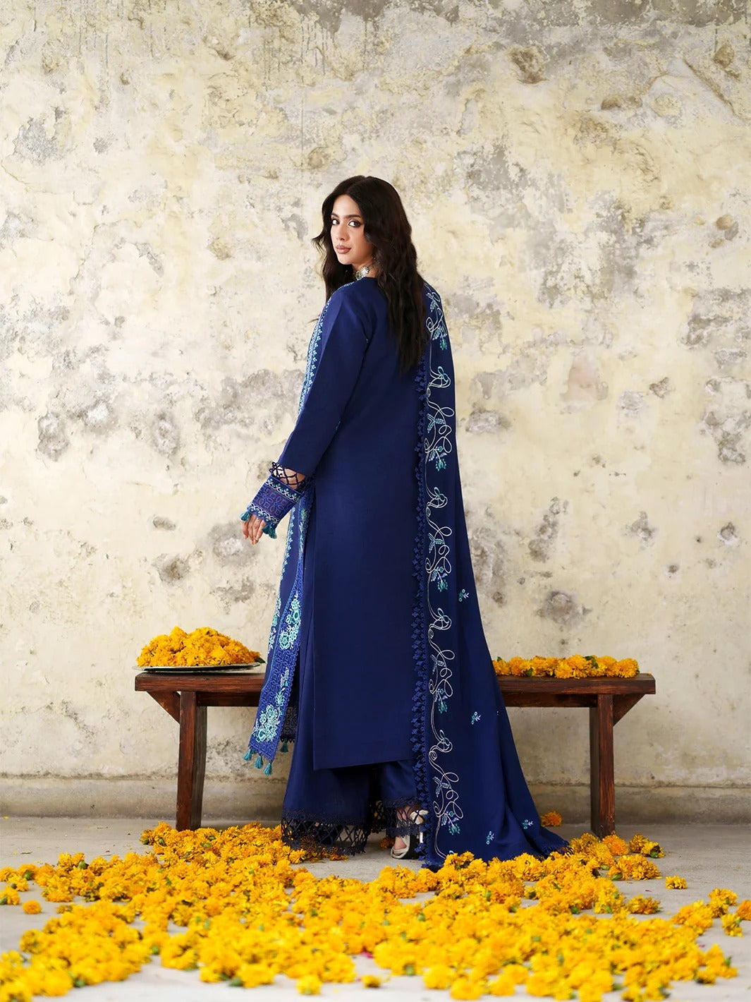 A woman wearing a deep blue traditional outfit with intricate white embroidery stands beside a wooden bench scattered with yellow marigold flowers, set against a textured beige wall.