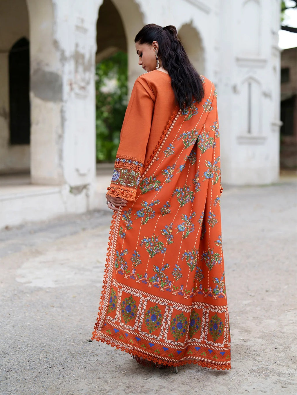 A woman wearing a vibrant orange traditional outfit with intricate floral embroidery and a matching dupatta, photographed from behind in an outdoor courtyard setting.