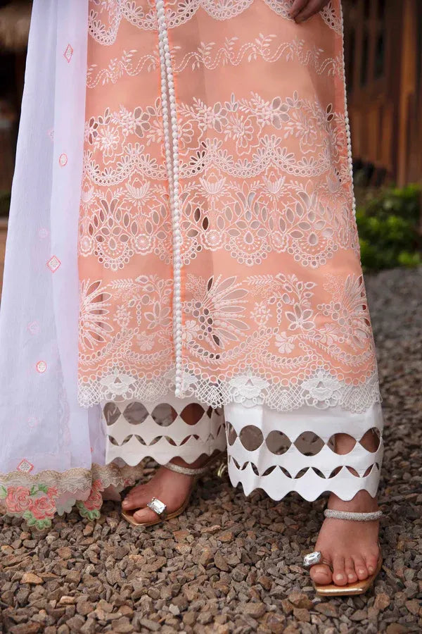 A close-up of a person wearing a peach and white embroidered traditional outfit with lace detailing, white patterned pants, and a sheer white dupatta, standing on a gravel surface.