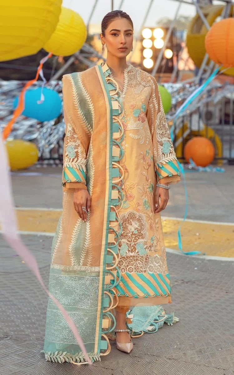 A woman models a traditional three-piece salwar kameez suit in beige and turquoise. The outfit features intricate gold embroidery, scalloped borders, and a matching turquoise dupatta, set against a festive outdoor background with colorful balloons.