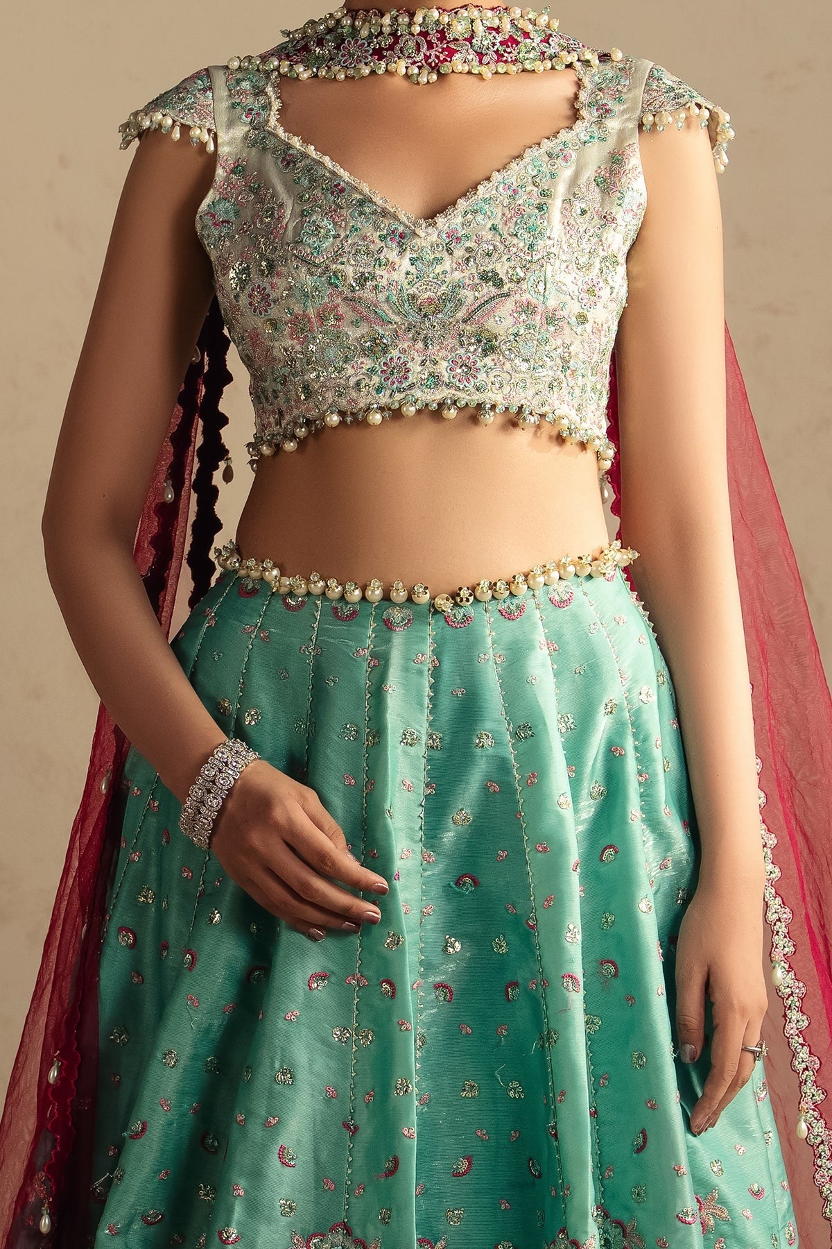 A close-up of a model wearing a traditional Indian lehenga choli set. The outfit features a mint green skirt with gold and pink floral embroidery, a matching embroidered crop top, and a pink dupatta draped over the shoulder.