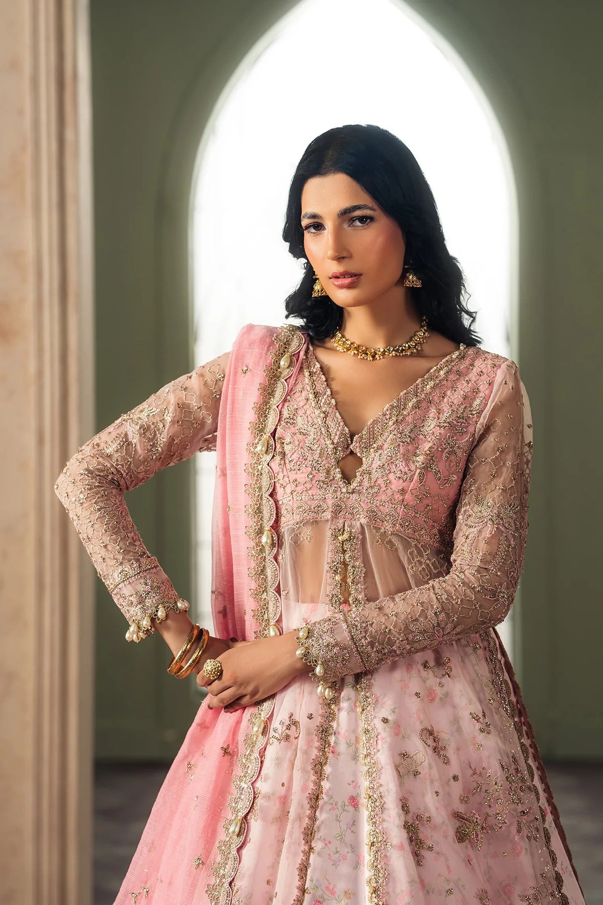 A woman modeling a pink traditional embroidered outfit with sheer long sleeves, floral detailing, and a matching pink dupatta, posed in front of an arched window.
