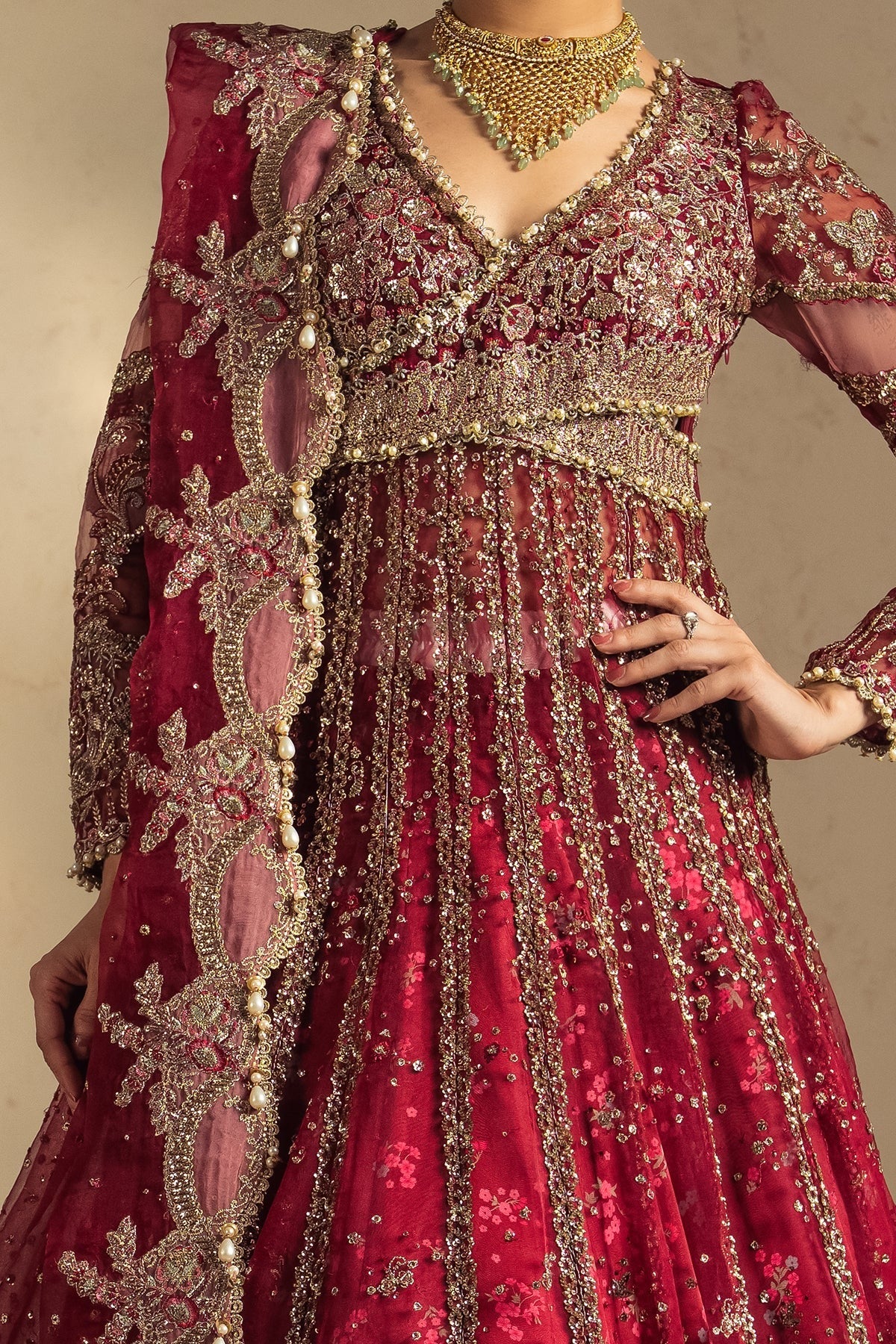A close-up of a heavily embellished traditional red and gold lehenga choli set with a matching embroidered dupatta, worn by a model against a neutral beige background.