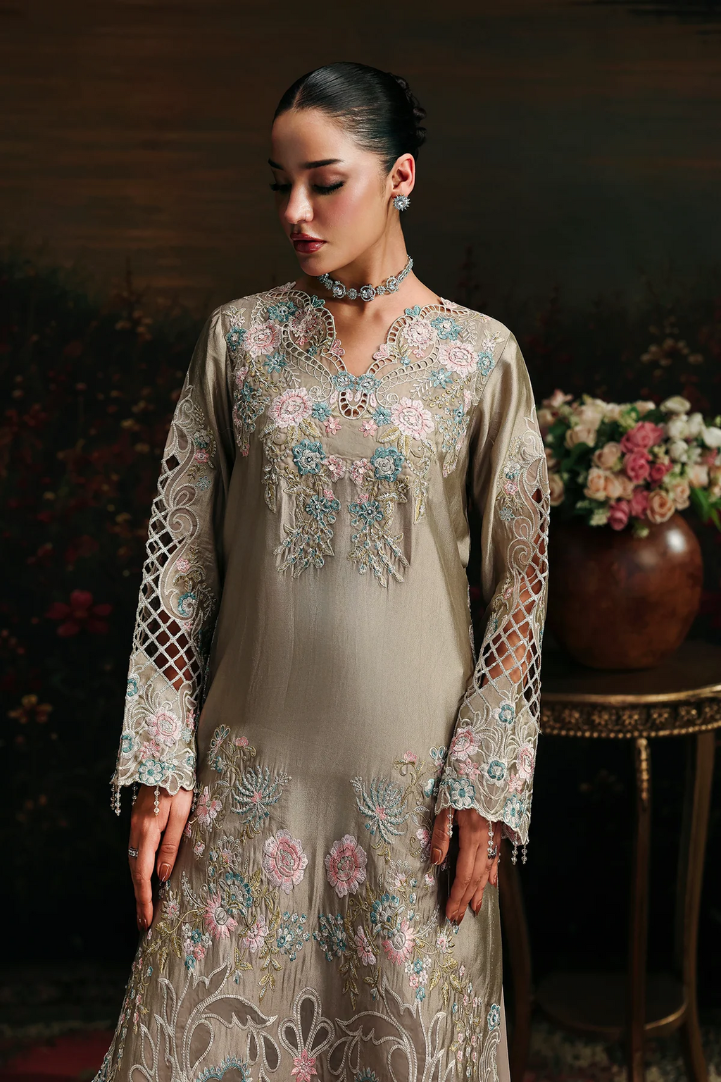 A woman models a traditional long-sleeve beige gown with intricate floral embroidery in pink, blue, and green, set against a dark, elegant background with a floral arrangement.