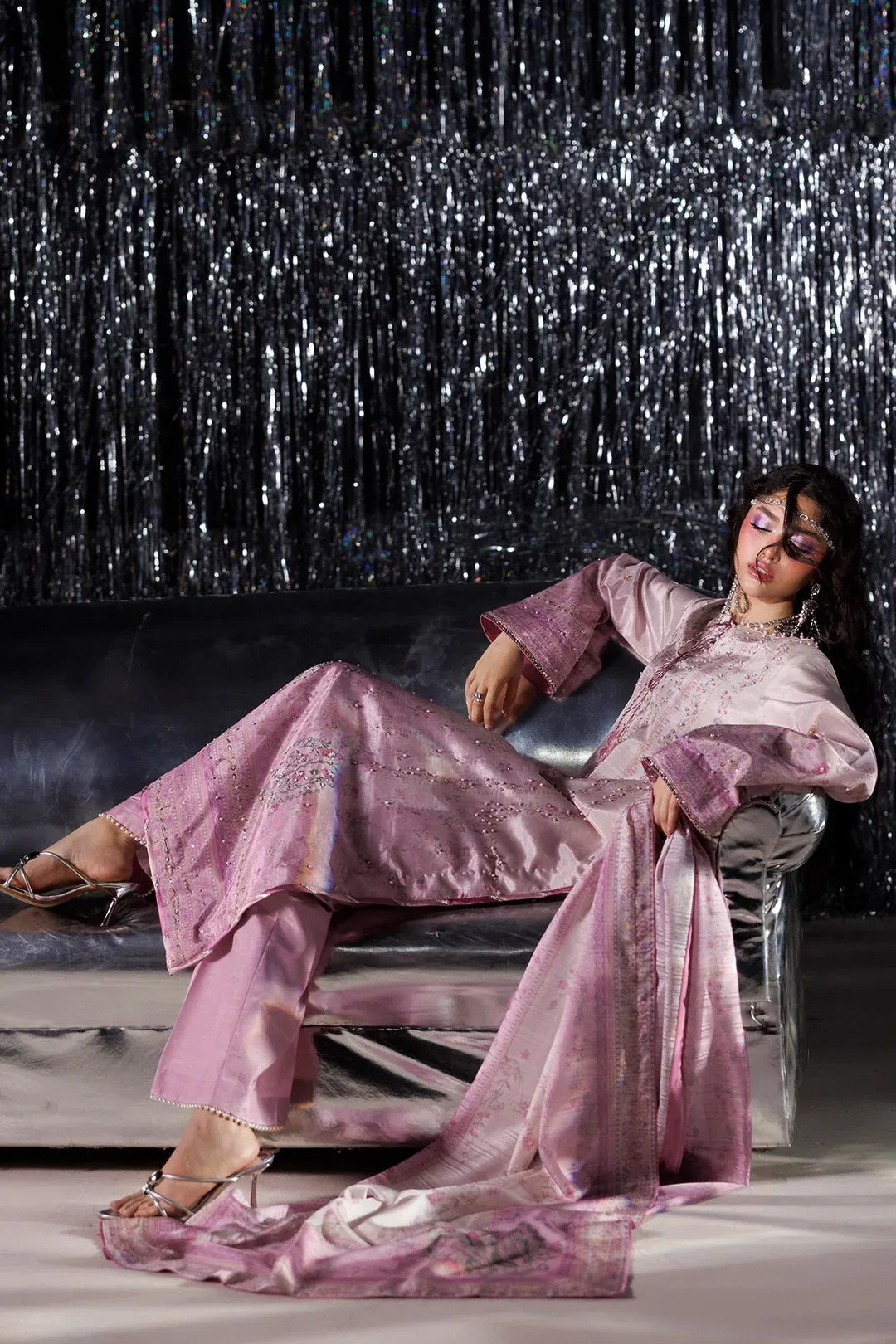 A woman reclines on a metallic sofa wearing a lavender-pink traditional outfit with matching trousers and a sheer draped scarf, posed against a black tinsel curtain backdrop.