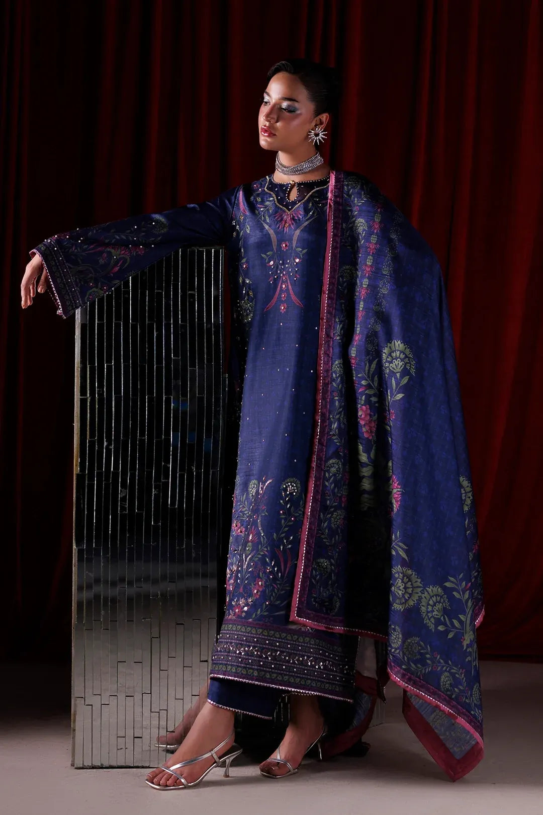 A model wears a traditional Indian salwar kameez suit in deep navy blue with intricate pink and gold floral embroidery, posed against a dark red curtain backdrop.