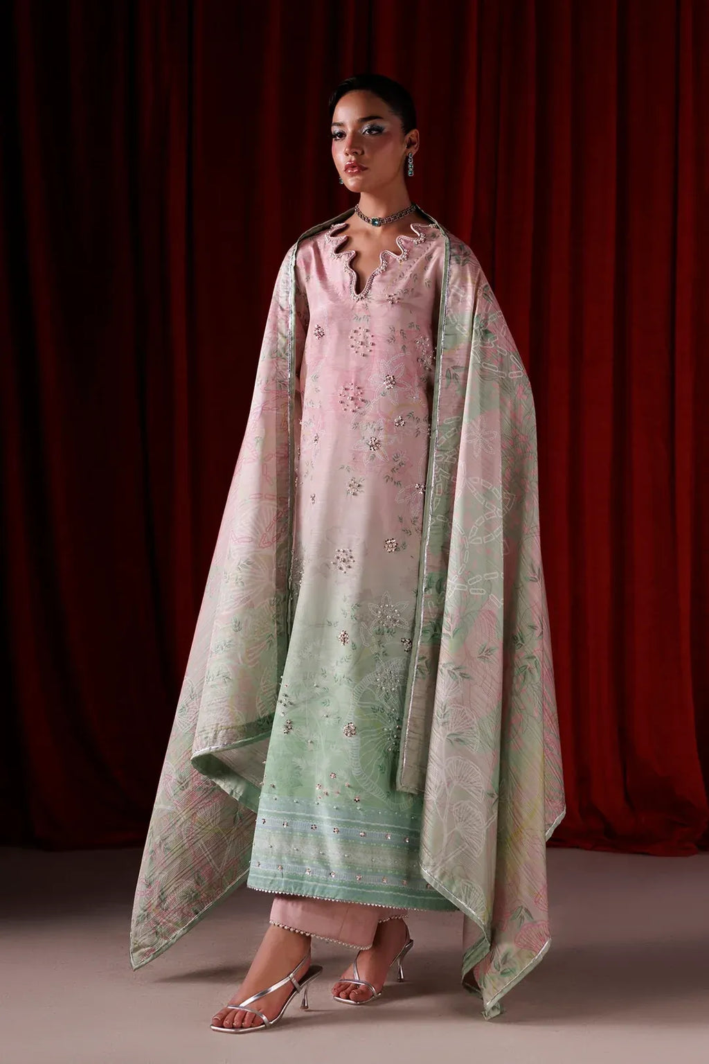 A model wearing a traditional South Asian outfit consisting of a light pink kurta with delicate floral embroidery, a matching light green dupatta with a floral pattern, and a pink shawl draped over her shoulders, set against a dark red curtain background.