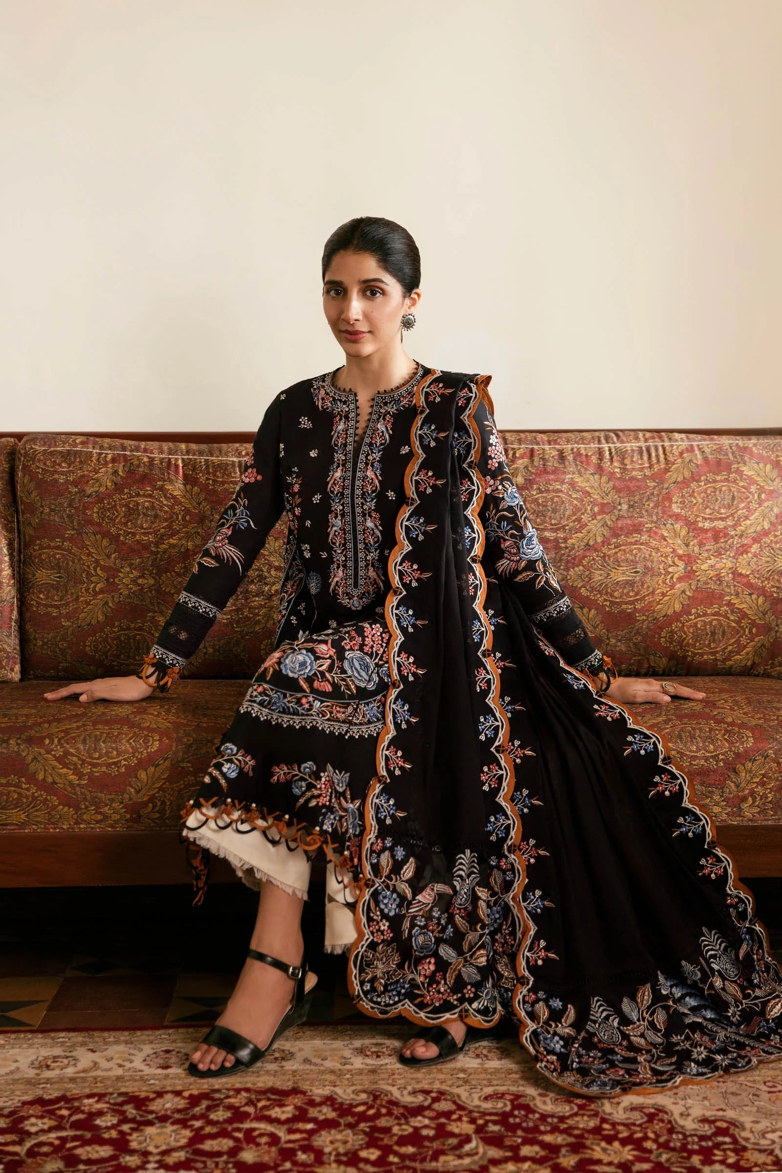 Woman in a black embroidered traditional outfit sitting on a patterned sofa.