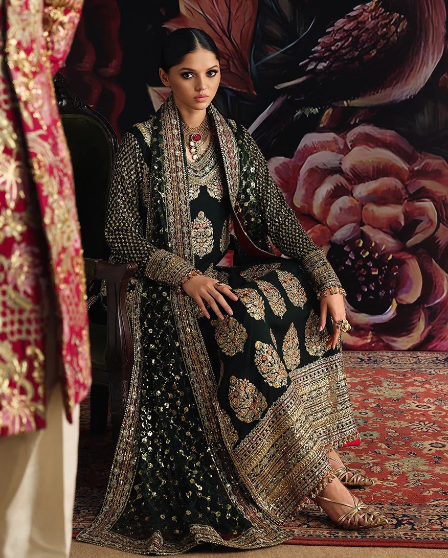 A woman seated indoors wearing an ornate black traditional outfit with extensive gold embroidery and mirror work, styled with layered jewelry and a patterned red rug. A large floral wall backdrop and part of another embroidered garment are visible.