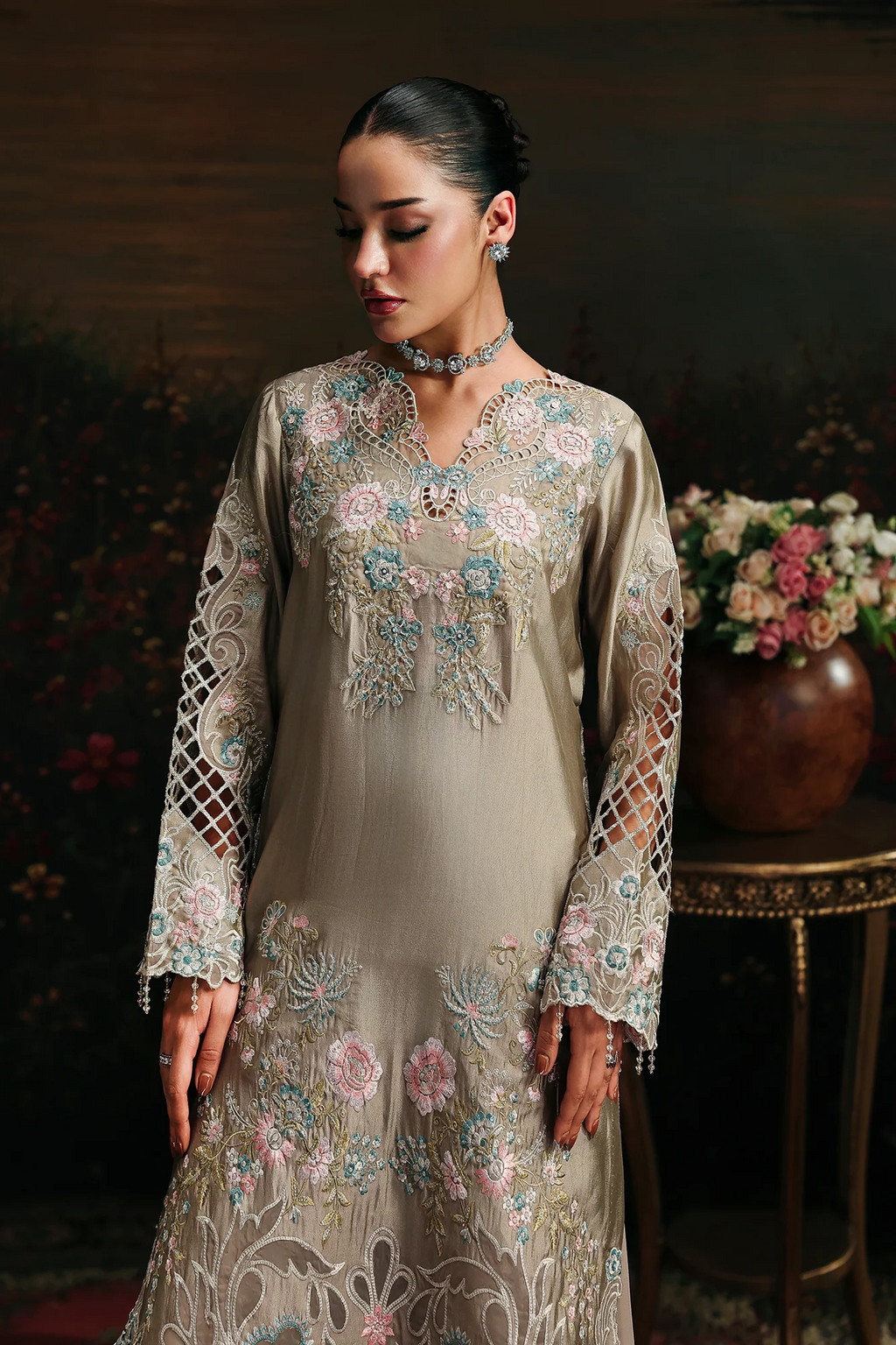 A woman wearing a beige embroidered traditional dress with floral patterns in pink, blue, and green, standing in a dimly lit indoor setting with a floral arrangement in the background.
