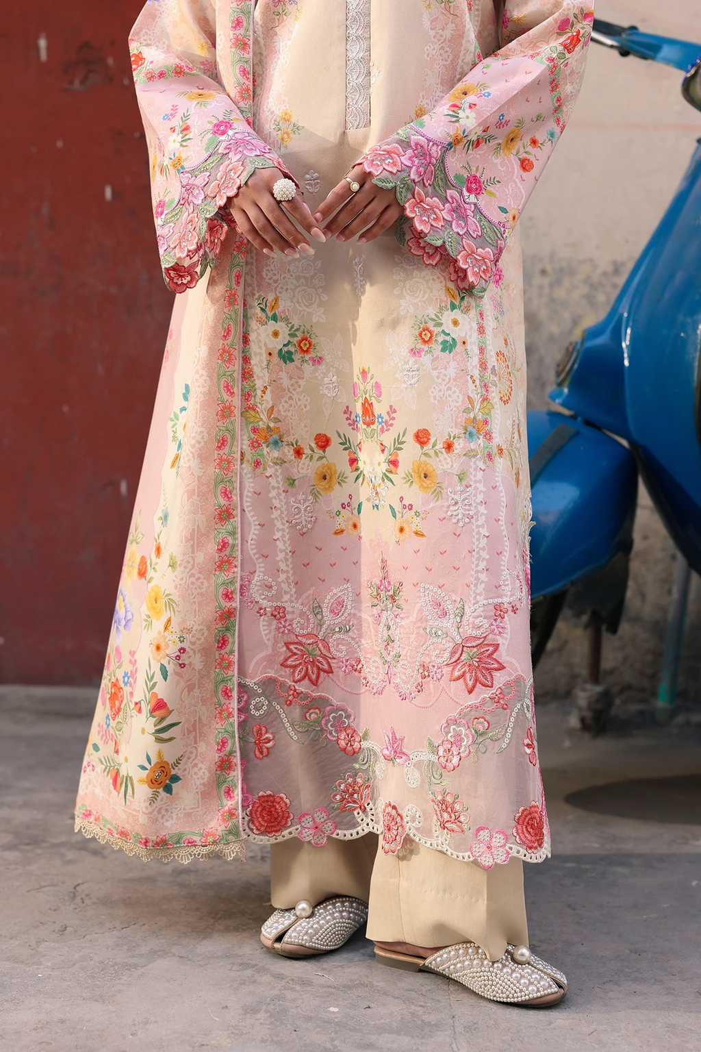 A person wearing a long, light pink embroidered tunic or caftan with floral patterns, paired with beige trousers and embellished flat shoes, standing outdoors next to a blue scooter.