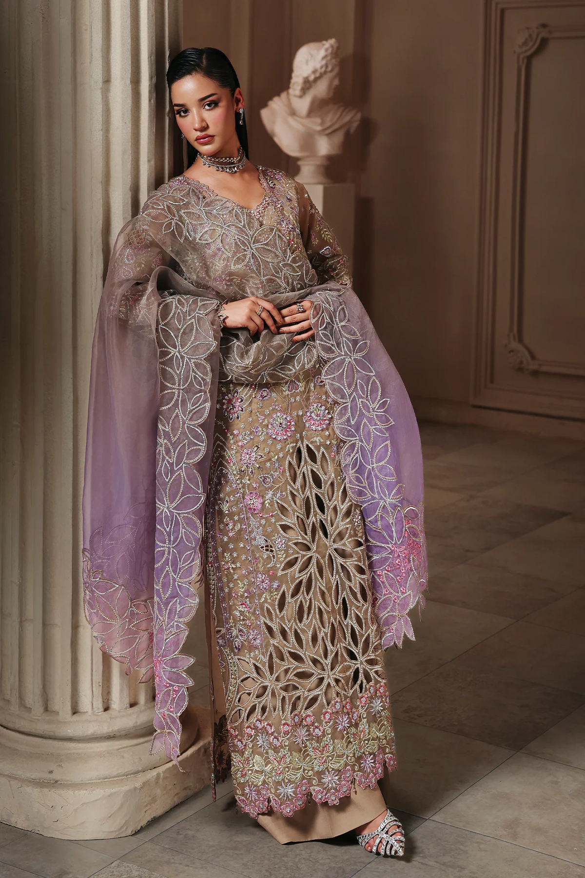 A woman models a heavily embroidered traditional outfit in an elegant indoor setting with a stone column and classical bust sculpture. The outfit features a beige base with intricate floral embroidery, a lavender sheer dupatta, and pink floral accents.