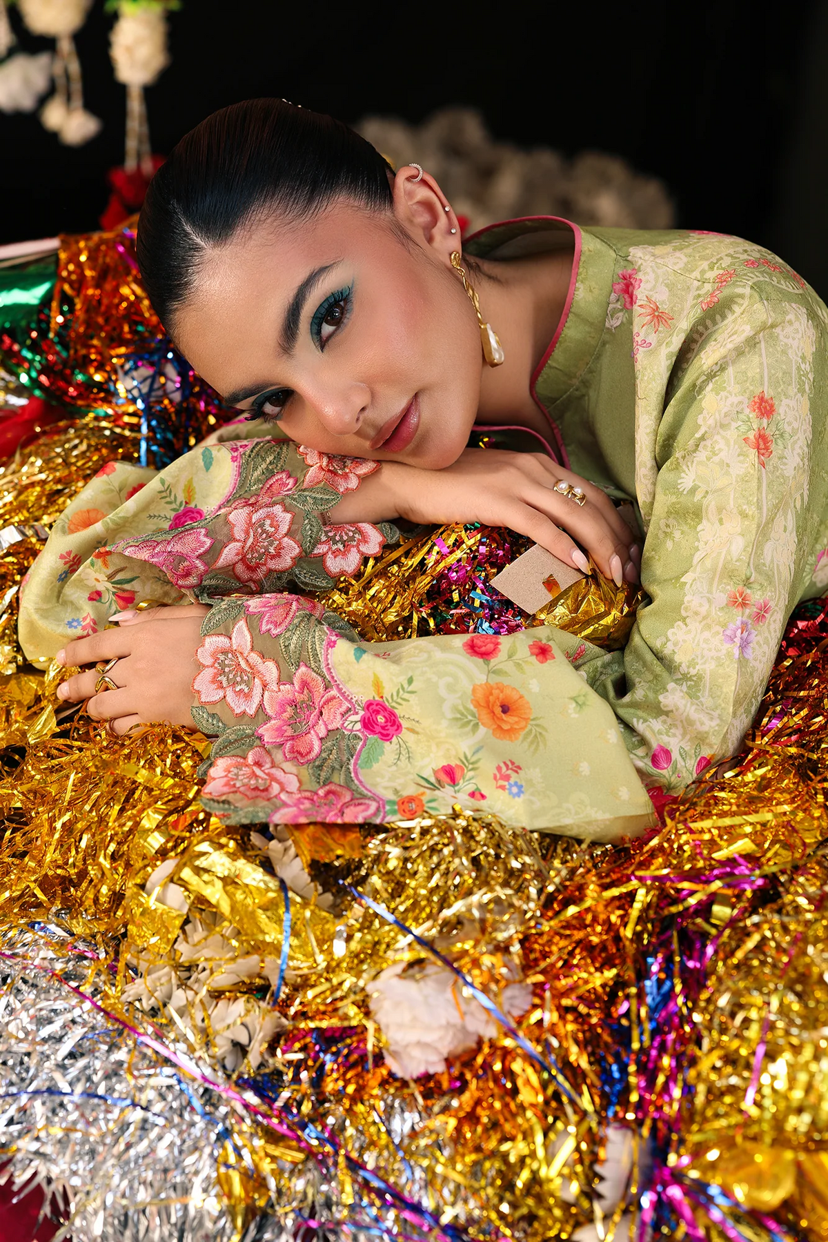 A woman in a light green floral traditional outfit rests on a bed of shiny gold and silver tinsel against a dark background.