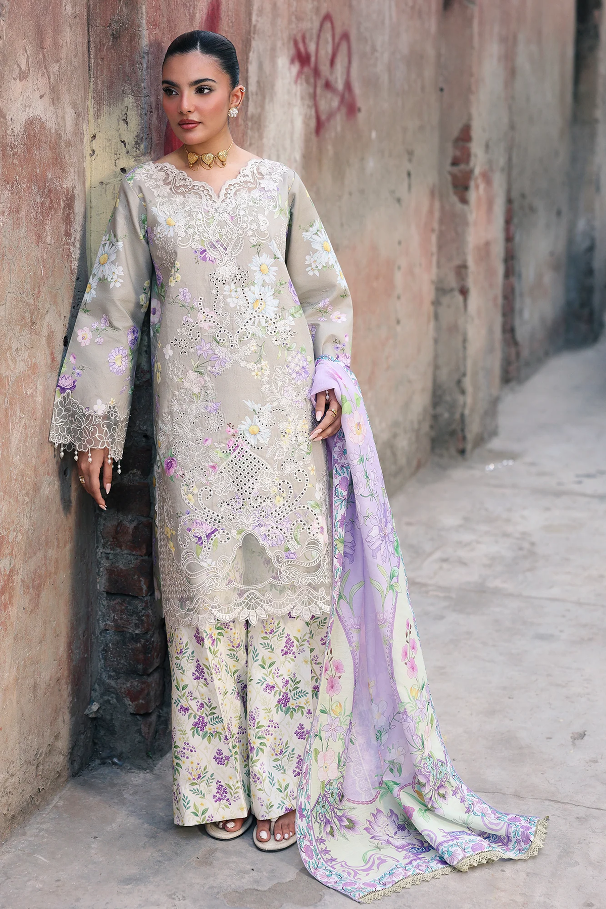 A woman models a traditional three-piece salwar kameez suit in a dusty alley. The set features a light beige embroidered kameez with a floral print and a matching dupatta in lavender, cream, and floral tones.