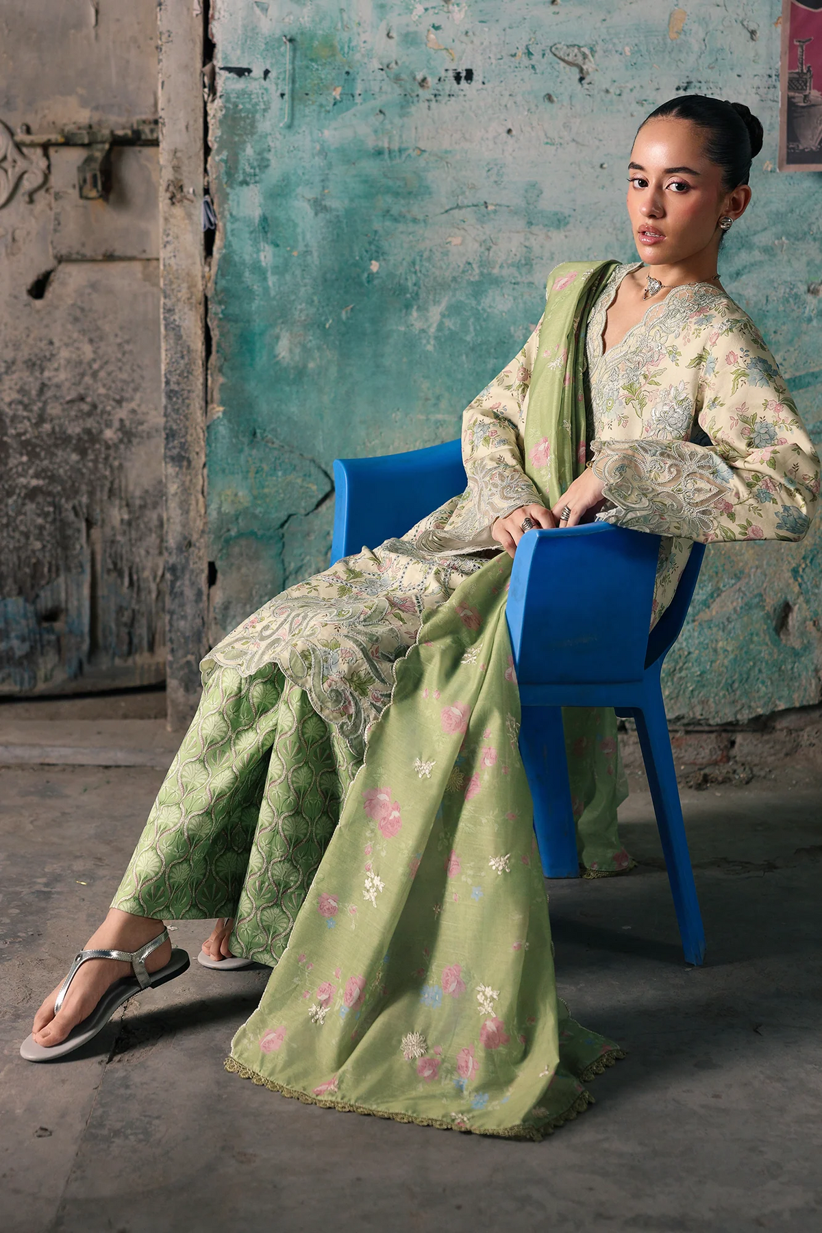 A woman models a light green floral print traditional South Asian outfit with matching pants and a dupatta, seated on a blue plastic chair against a rustic, weathered teal wall.