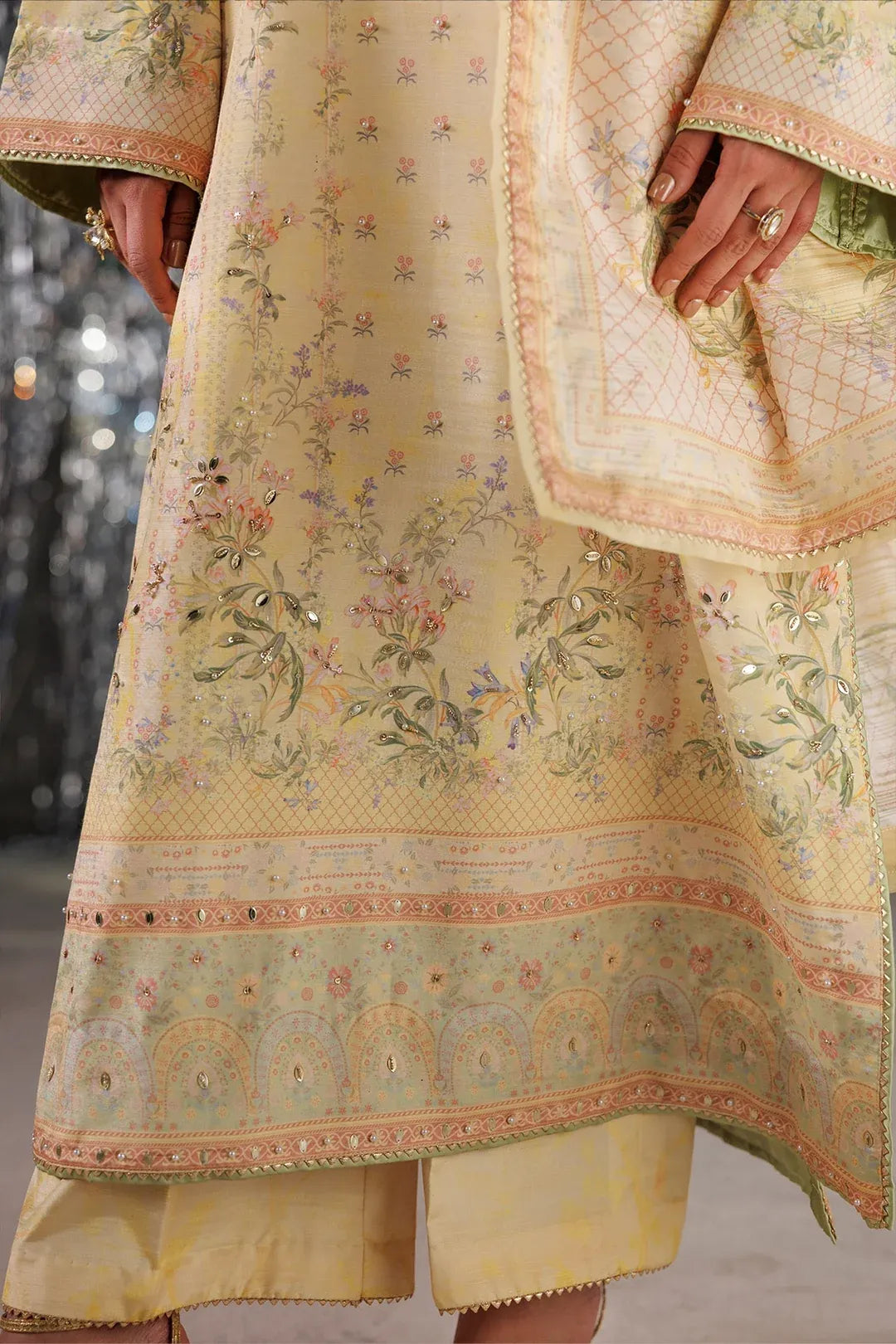 A close-up of a traditional embroidered outfit in pale yellow and beige, featuring floral patterns, intricate borders, and a matching dupatta held by a model's hand.