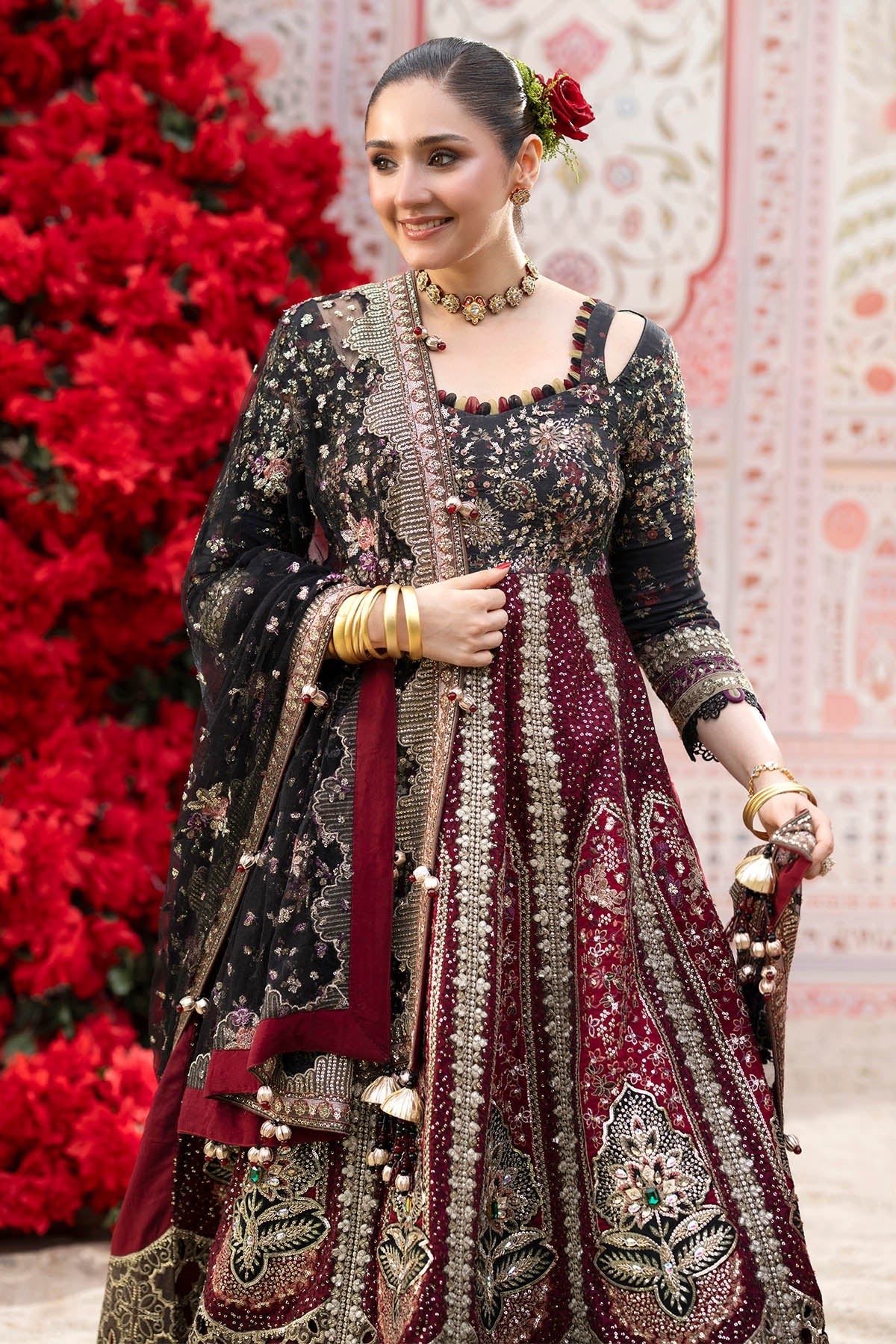 A woman models an ornate traditional Indian anarkali suit with heavy embroidery. The outfit features a black upper section with gold embroidery and a deep maroon lower skirt with intricate gold and silver embellishments.