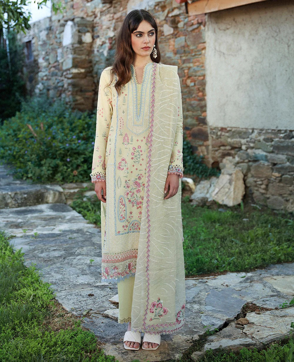 Woman in a light-colored embroidered traditional outfit standing outdoors with a stone wall and greenery in the background.