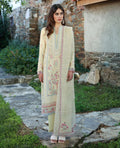 Woman in a light-colored embroidered traditional outfit standing outdoors with a stone wall and greenery in the background.