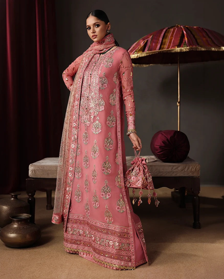 A woman models a traditional pink embroidered outfit with a matching dupatta and a small matching embroidered pouch bag, posed in an ornate studio setting with a striped umbrella, cushions, and decorative pots.