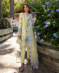 Woman in a light yellow traditional outfit with floral patterns standing near a stone wall and plants.