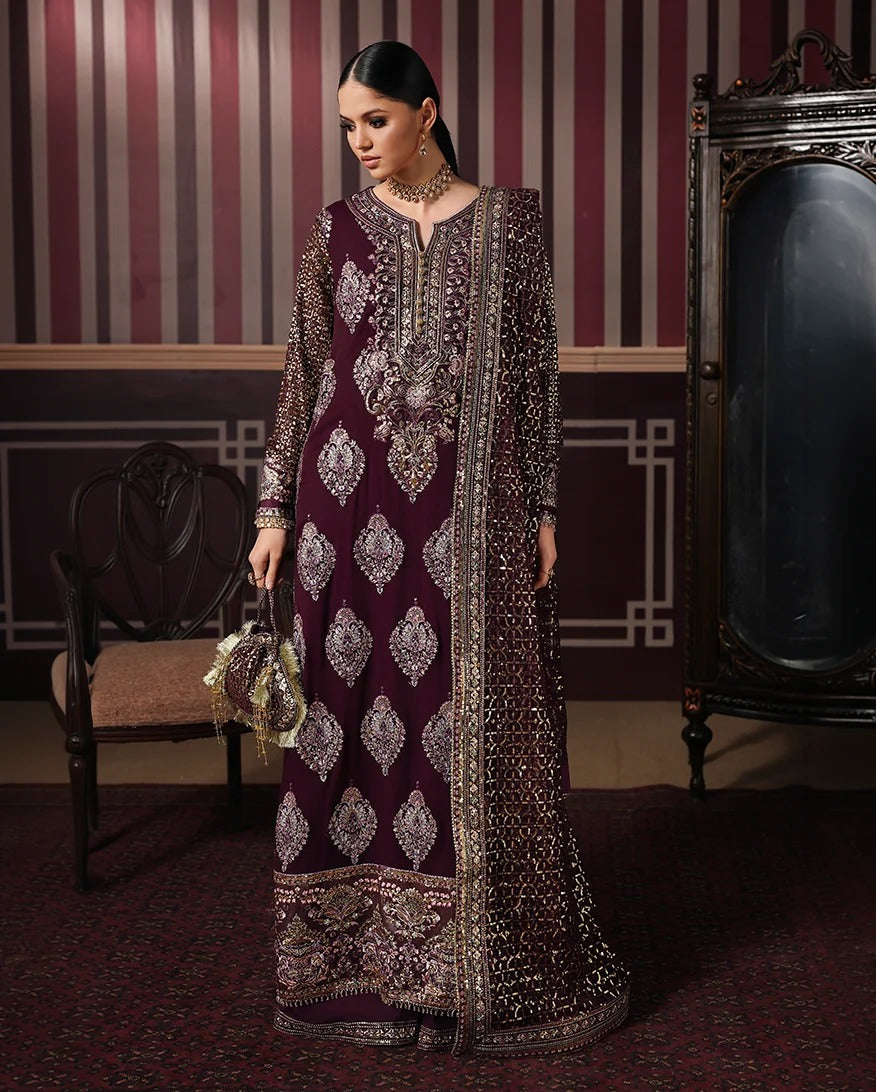 A woman models a traditional long gown in deep maroon with intricate silver and gold embroidery, standing in a vintage room with striped wallpaper, a mirror, and a chair.