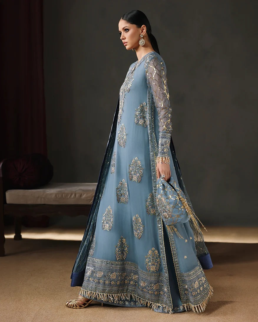 A woman models a traditional blue anarkali gown with intricate gold embroidery, paired with a matching sheer dupatta and clutch bag, set against a dark, elegant indoor background.