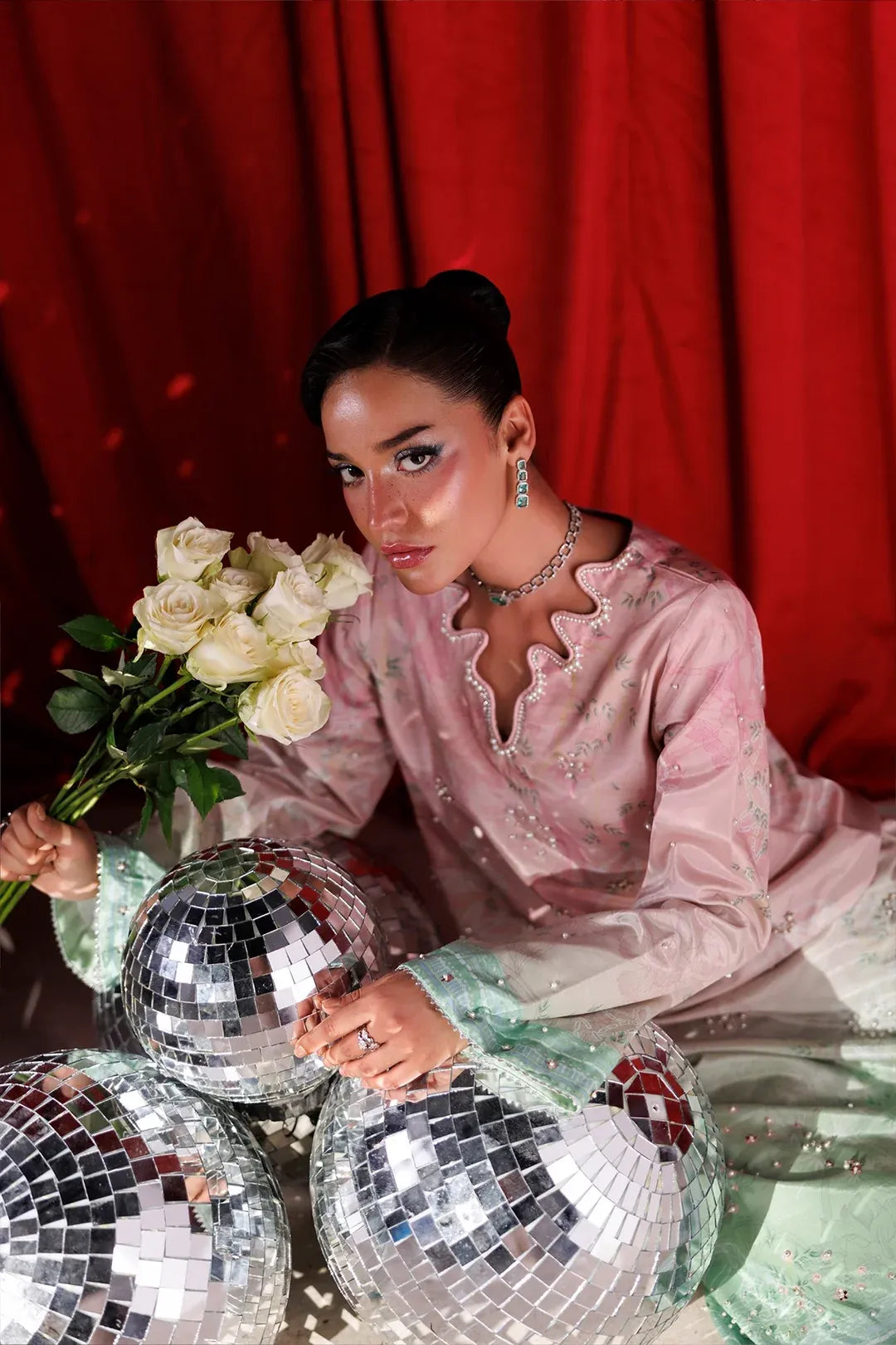 A woman in a pale pink embroidered traditional outfit sits in front of a red curtain, holding a bouquet of white roses and surrounded by mirrored disco balls.