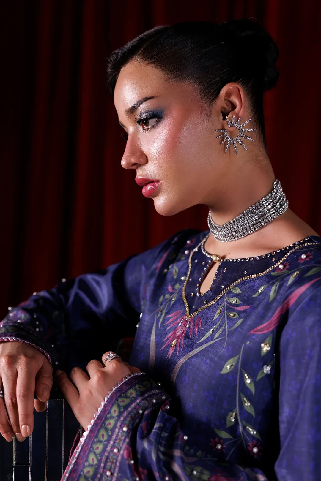 A woman in profile wearing a dark purple embroidered traditional outfit with metallic embellishments, a silver choker necklace, and a star-shaped earring, posed against a dark red curtain background.