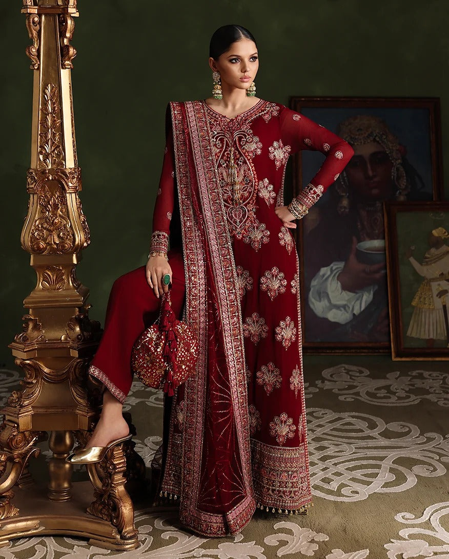 A woman models a traditional deep red embroidered ethnic outfit with matching pants and a draped scarf, posed next to an ornate gold column in a dark, elegant interior setting.