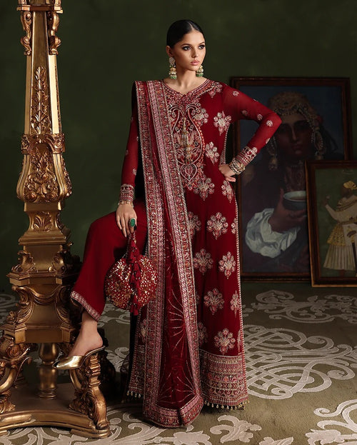 A woman models a traditional deep red embroidered ethnic outfit with matching pants and a draped scarf, posed next to an ornate gold column in a dark, elegant interior setting.