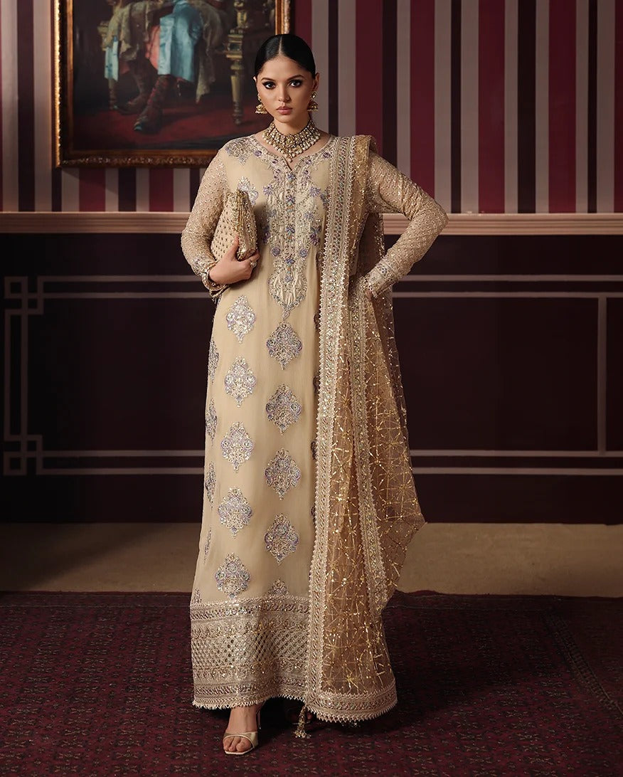 A woman models a traditional beige Anarkali suit with intricate silver and gold embroidery, paired with a matching sheer dupatta, in an ornate room with striped walls and a framed painting.