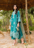 A woman models a teal green ethnic three-piece suit with intricate white embroidery, featuring a long kurta, matching pants, and a sheer dupatta, set against a tropical outdoor patio background.