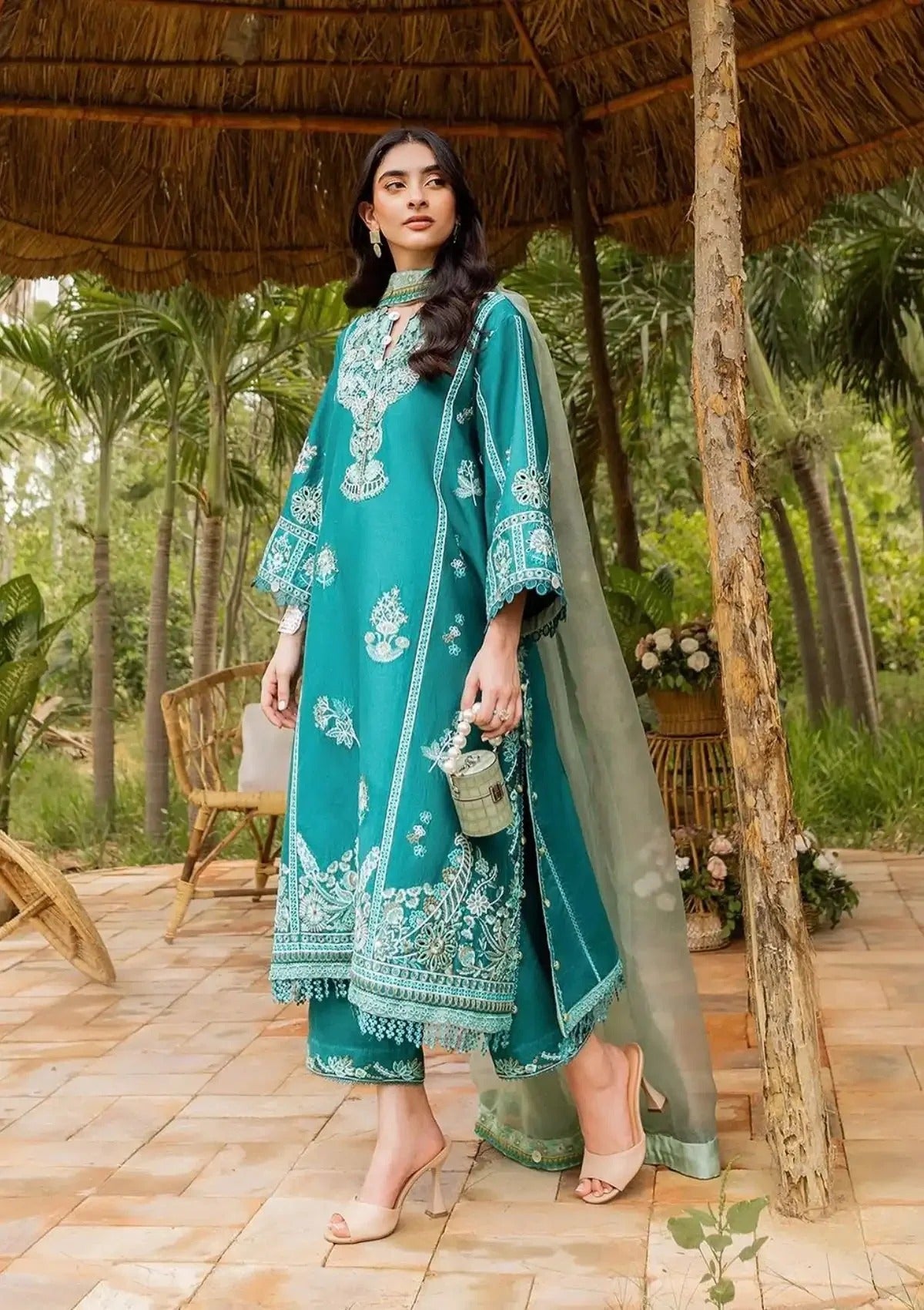 A woman models a teal green ethnic three-piece suit with intricate white embroidery, featuring a long kurta, matching pants, and a sheer dupatta, set against a tropical outdoor patio background.