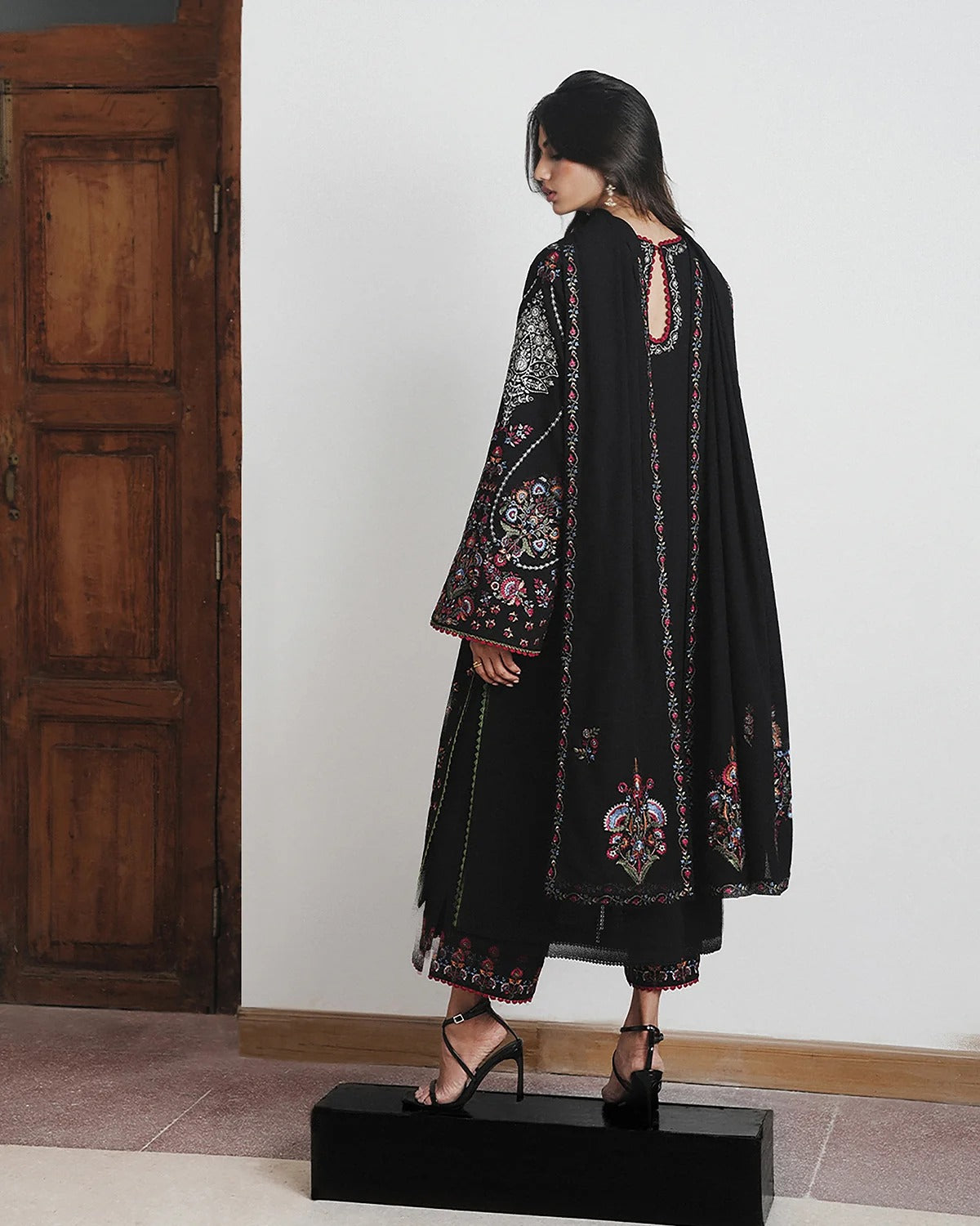 A woman modeling a black traditional outfit with colorful floral embroidery, standing on a black platform in front of a white wall and wooden door.