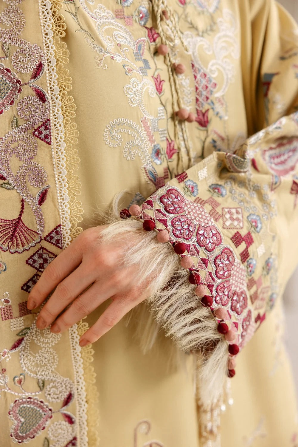 Close-up of a cream-colored embroidered traditional robe with intricate floral and paisley patterns in pink, purple, and white, featuring a decorative sleeve with faux fur trim and a hand resting on the fabric.