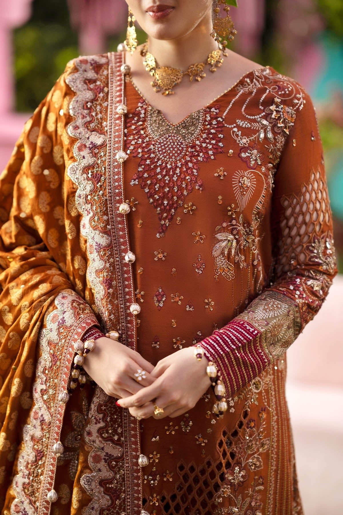 A close-up of a woman wearing an ornate rust-orange embroidered traditional outfit with a matching draped scarf, heavy metallic embroidery, and gold jewelry against a softly blurred pink background.