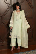 A woman models a light mint green traditional three-piece suit with intricate white floral embroidery and a matching sheer dupatta, standing in front of a dark wood paneled wall.