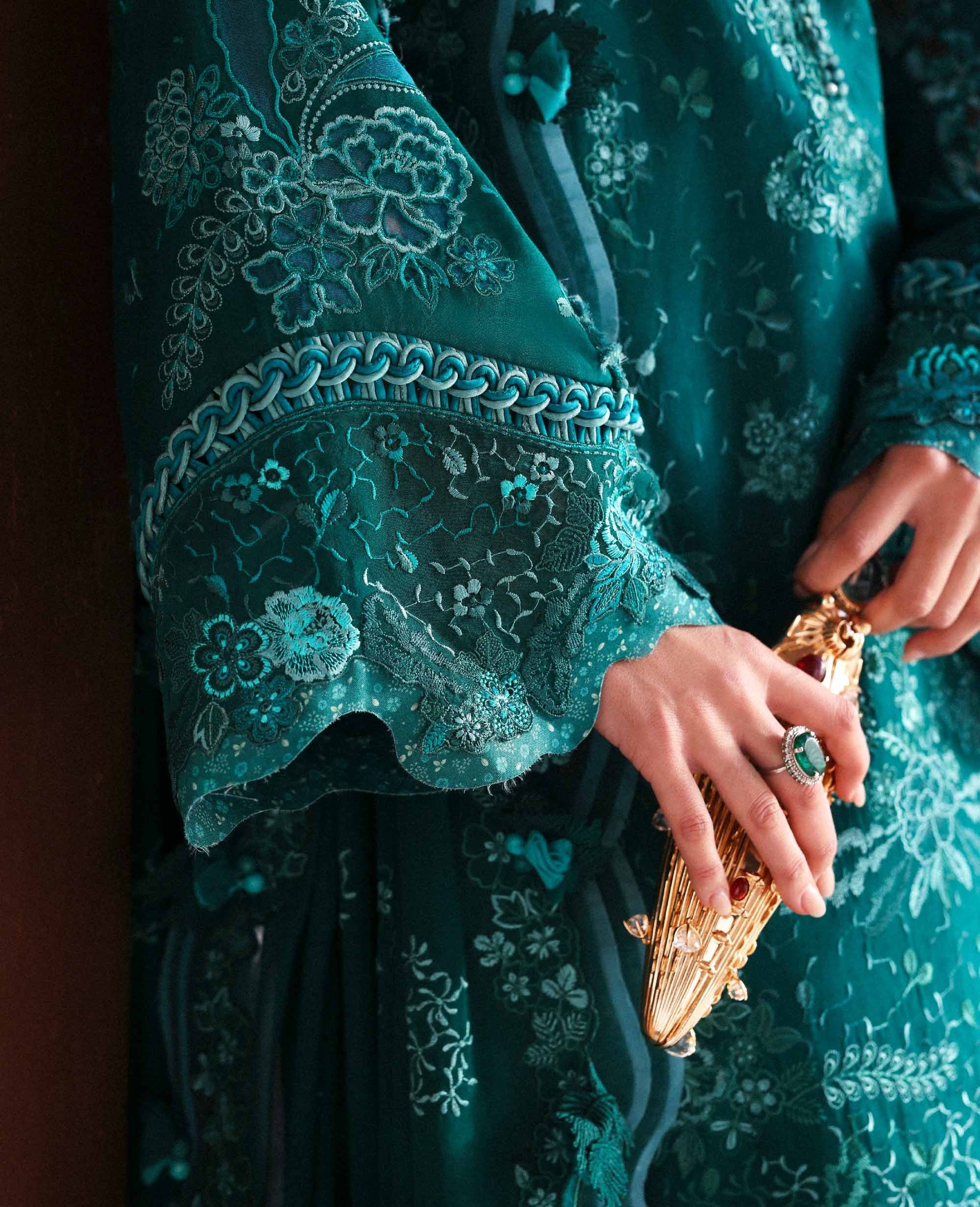 Teal embroidered garment with a hand holding a gold clutch.