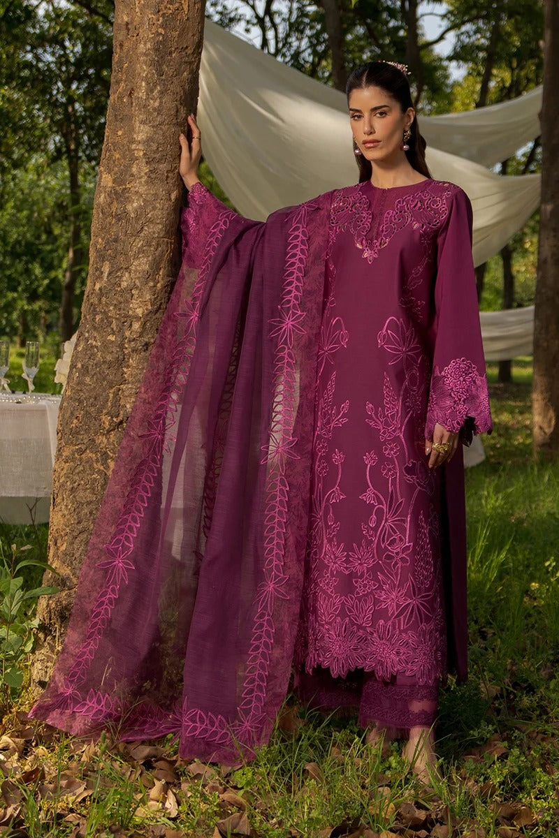 A woman wearing a deep purple embroidered traditional outfit with a matching sheer dupatta, standing outdoors next to a tree in a garden setting.